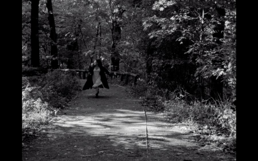 Refracted — still: black-and-white image of a woman running down a forest path, coat billowing